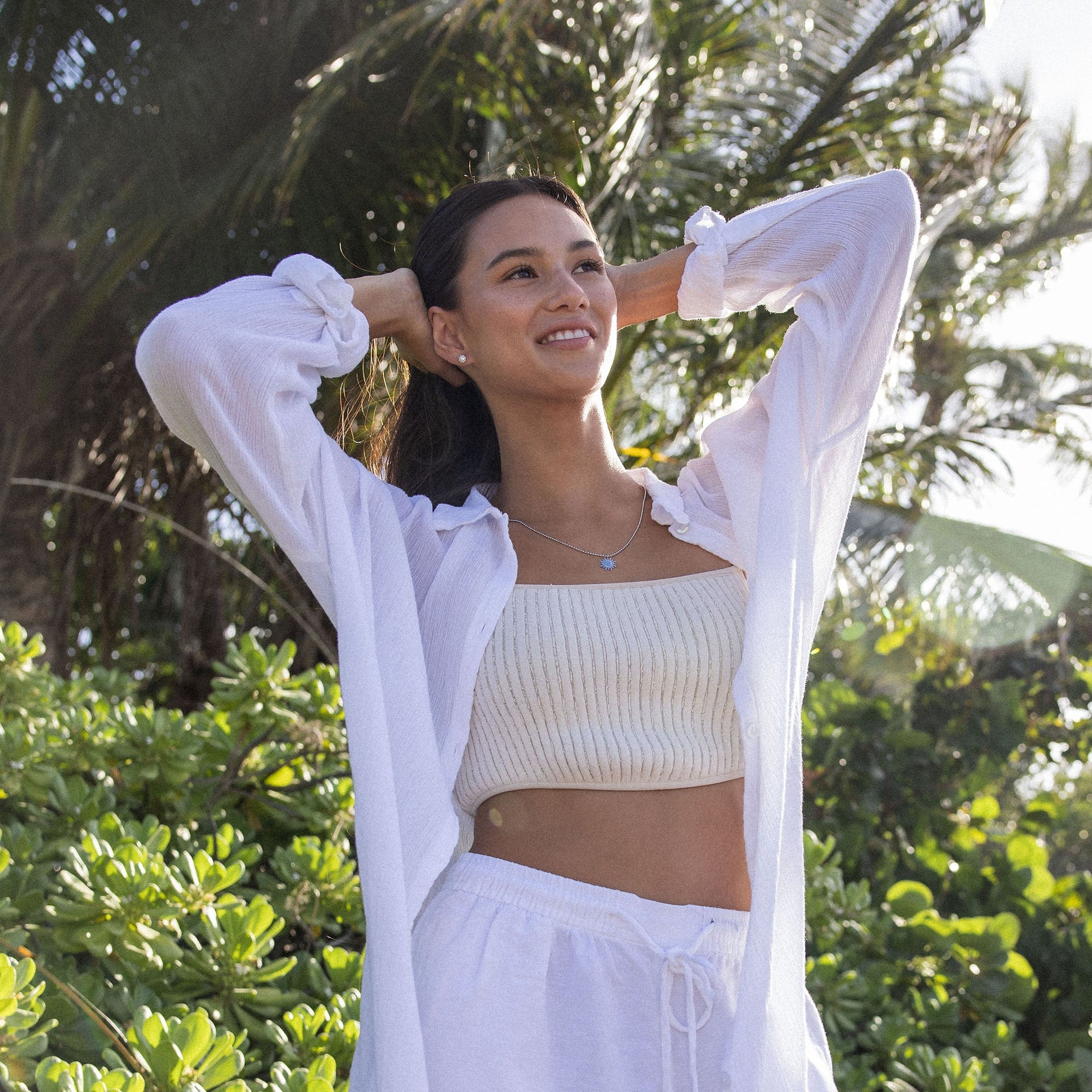 Feminine, weiße PURELEI Bluse im Sonnenlicht, umgeben von tropischem Grün.