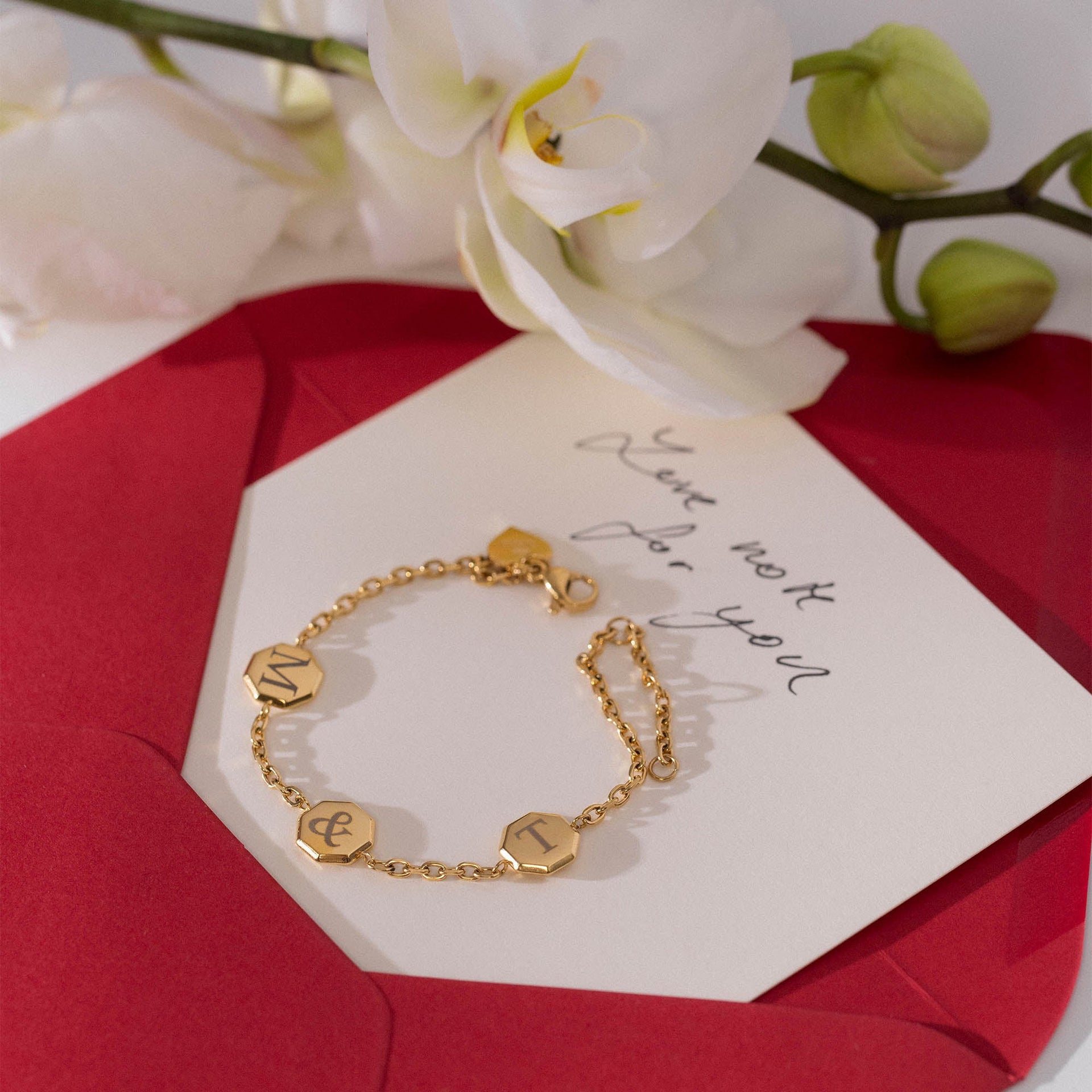 Goldenes Armband mit Buchstabenanhängern auf rotem Umschlag, PURELEI inspiriert.